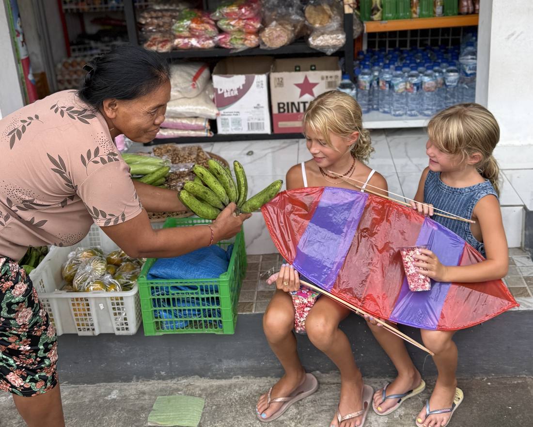 Een Balinese winkelierster geeft twee meisjes met een vlieger een tros bananen aan, Bali, Indonesië