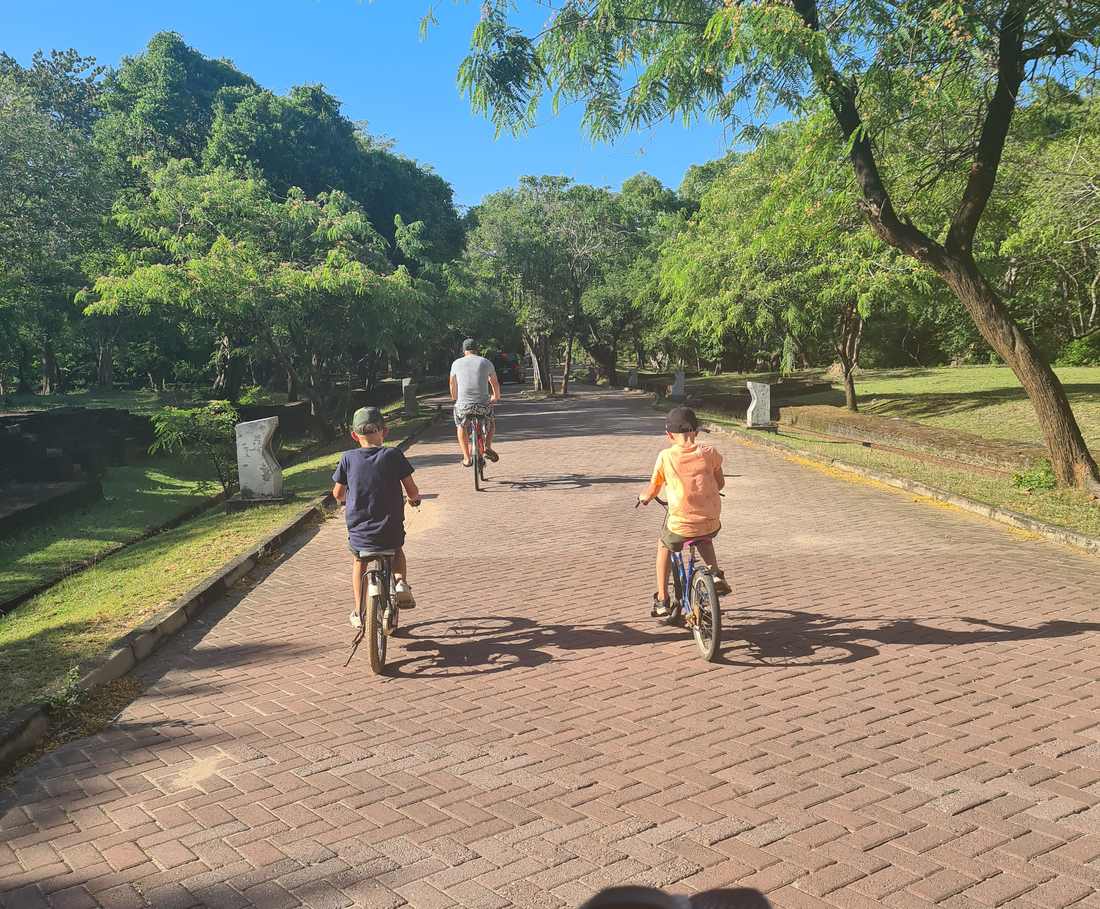 Een vader en twee kinderen fietsen over het rode klinkerpad tussen de ruines van Polonnaruwa, Sri Lanka