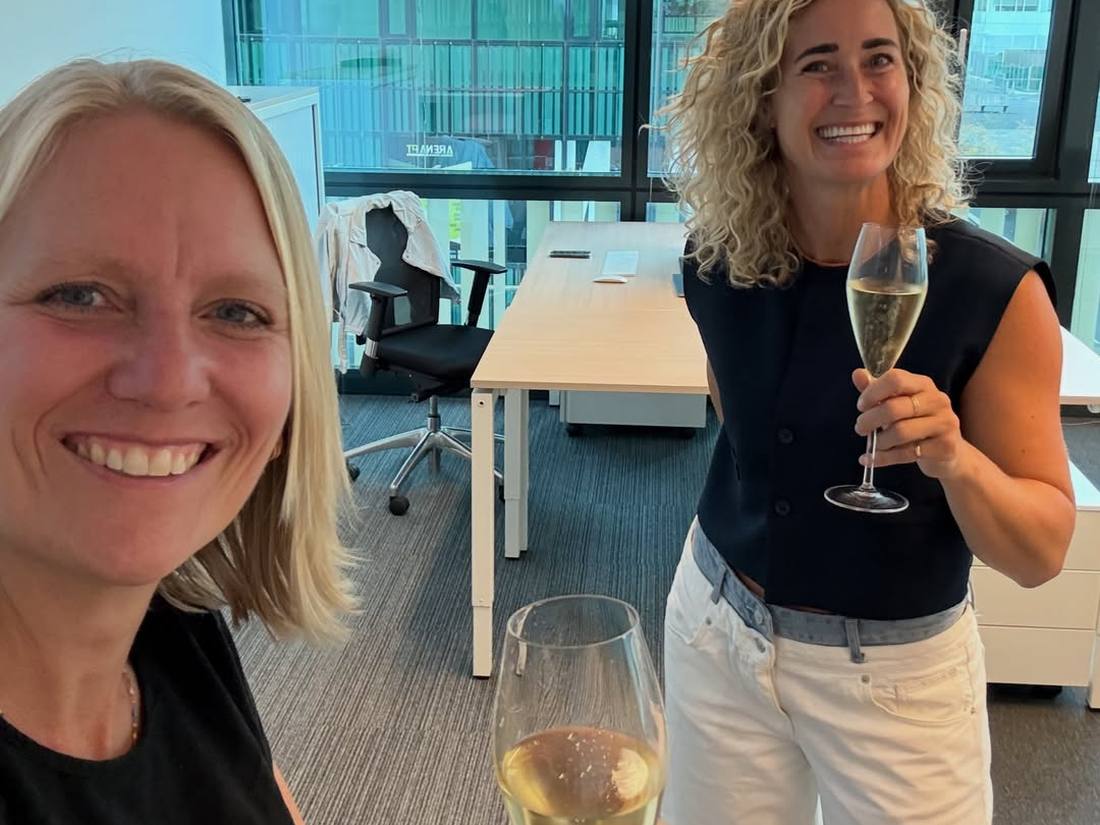 Nynke en Ilse proosten met een glas champagne in hun nieuwe kantoor in Amsterdam Zuidoost, allebei lachend in beeld.