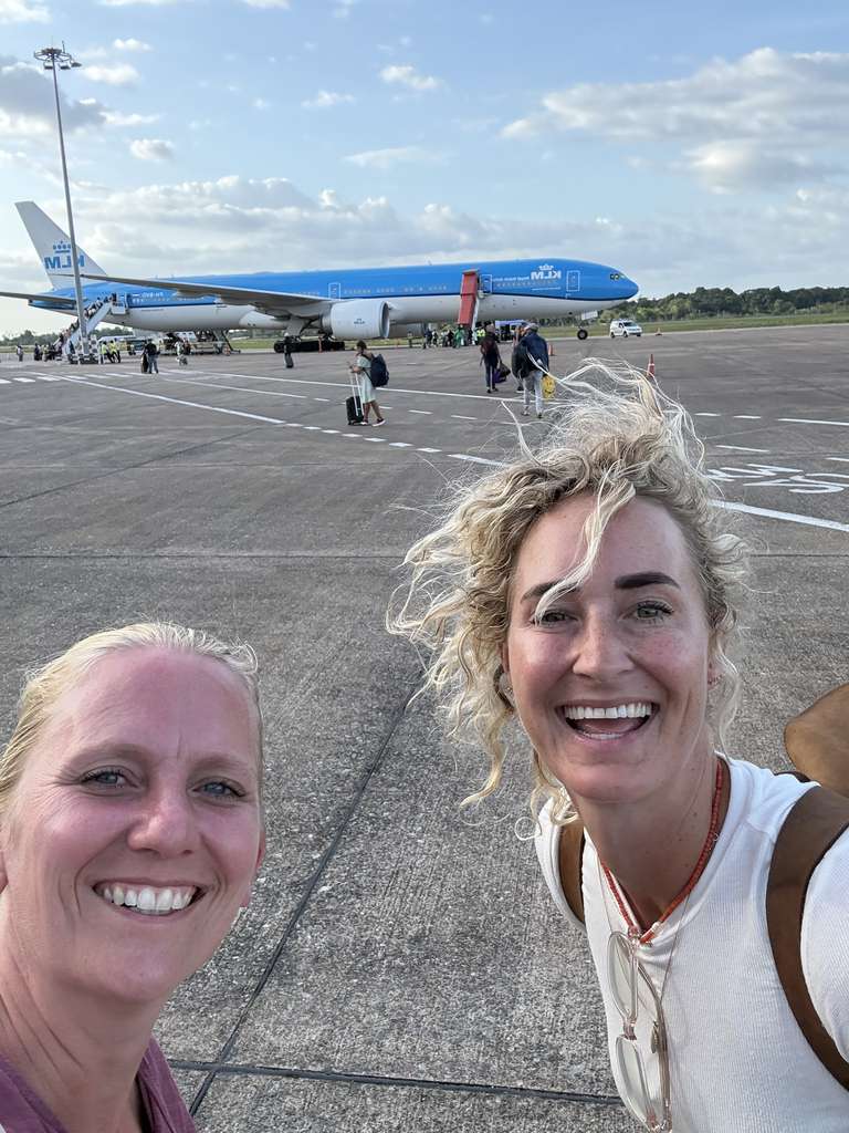 Nynke en Ilse lachend op het platform van het vliegveld, met het vliegtuig achter zich