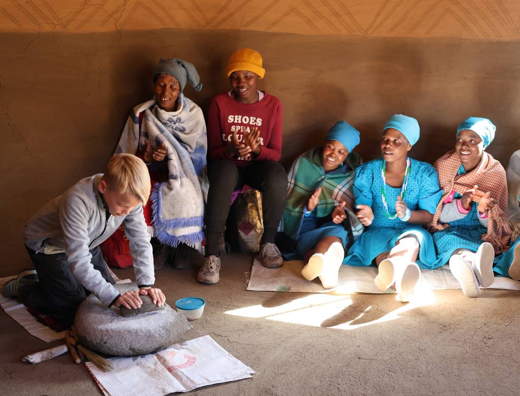 Jongen maalt mais op een steen terwijl vrouwen in klederdracht meeklappen, Zuid-Afrika