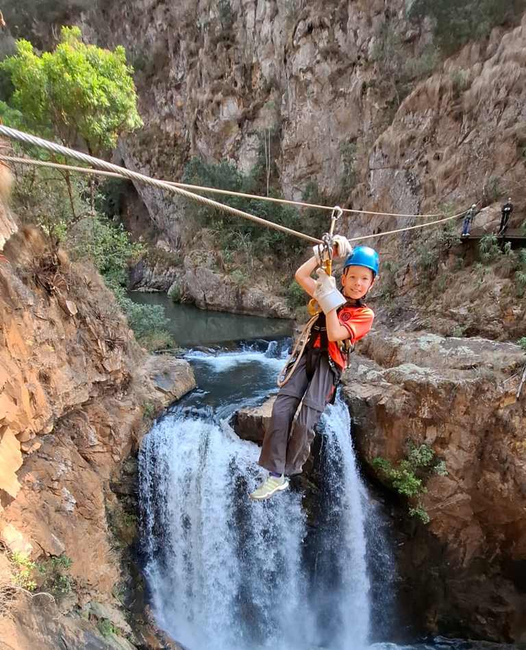 Kind zweeft met helm aan een zipline over een waterval in een kloof, Zuid-Afrika